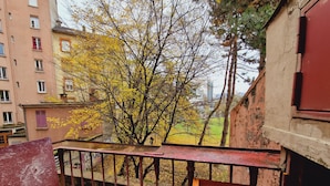 A balcony with a metal railing overlooking a tree with yellow leaves, a building with a red door, and a view of a park.