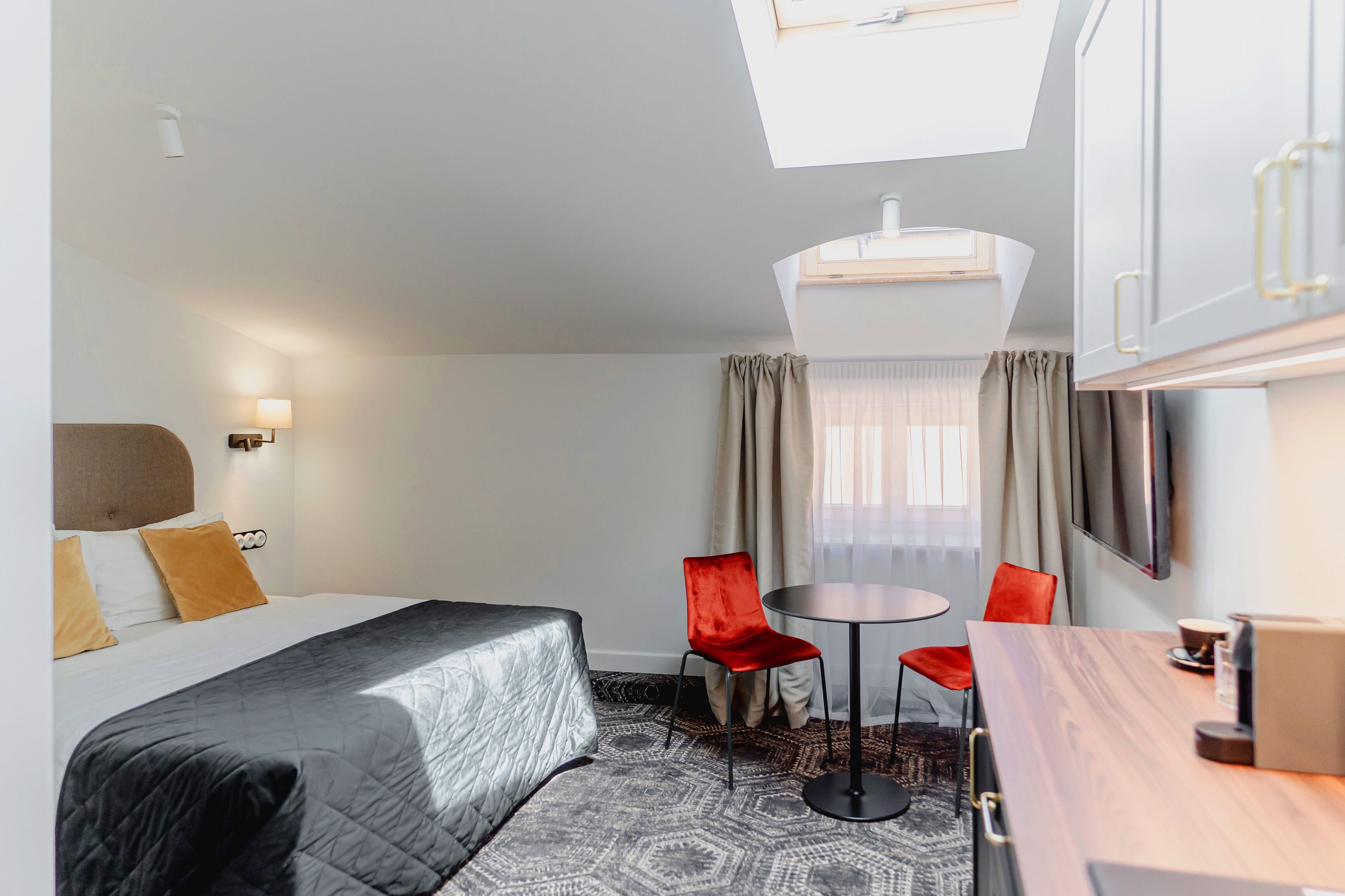 A hotel room with a bed, a desk, two chairs, a TV, and a window with curtains.