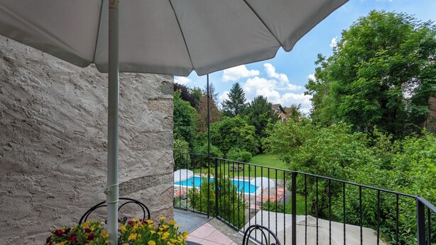 A balcony with a view of a swimming pool, surrounded by greenery and a stone wall.