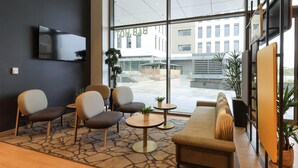 A modern interior with a sofa, chairs, a TV, and large windows offering a view of buildings outside. - B&B HOTEL Dijon Valmy Toison d'Or (Dijon)