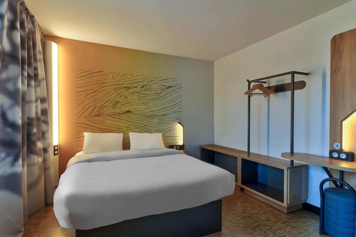 A hotel room with a bed, bedside tables, a desk, a coat rack, and a wall-mounted artwork.