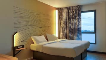 A modern hotel room with a large bed, a bedside table, a wall-mounted light, and a window with curtains.