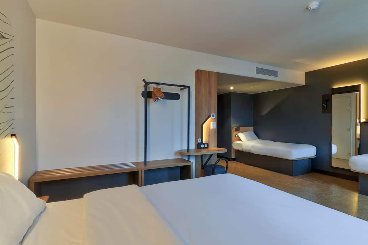 A hotel room with a large bed, a bedside table, a small table with a lamp, a bench, and a mirror.