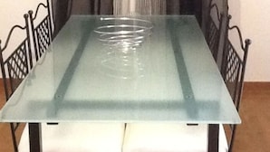 Une table à manger avec un plateau en verre et des chaises en métal, placée devant une fenêtre ornée d’un rideau léger et transparent.