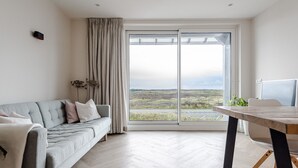 A modern living room with a sofa, a dining table, and a large window offering a view of a landscape. - Dunefront Apartment - A perfect getaway location! (Zandvoort)