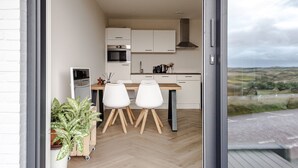 A modern kitchen with a dining area, featuring a wooden table and white chairs, a built-in oven, and a view of the countryside through a large window. - Dunefront Apartment - A perfect getaway location! (Zandvoort)