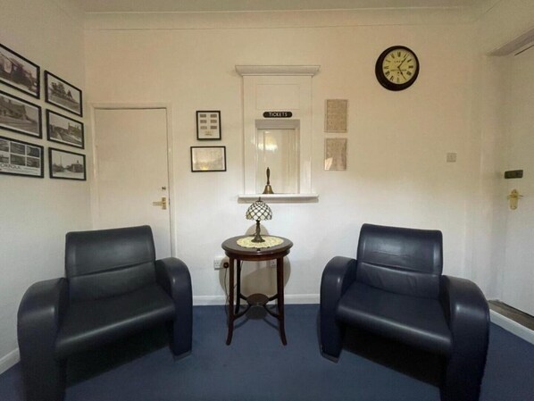 A room with two black leather chairs, a small round table with a lamp, a clock, and a door with a 'Tickets' sign above it.