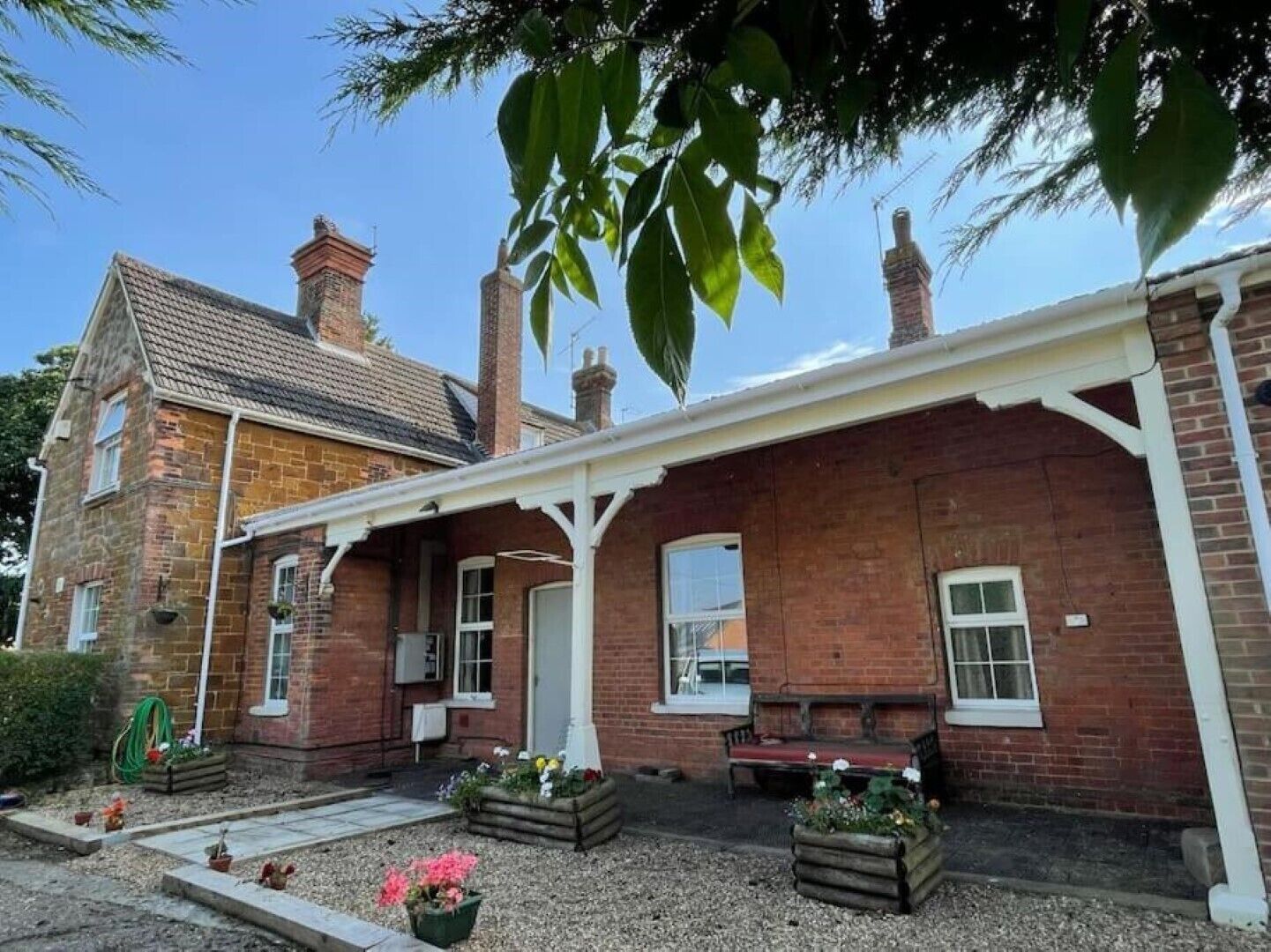 A two-story brick house with a covered porch, a garden with potted plants, and a bench.