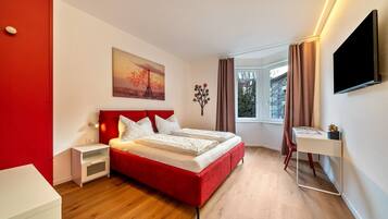 Una camera d'albergo moderna con un letto rosso, una scrivania bianca, una televisione a schermo piatto e un quadro raffigurante un tramonto.