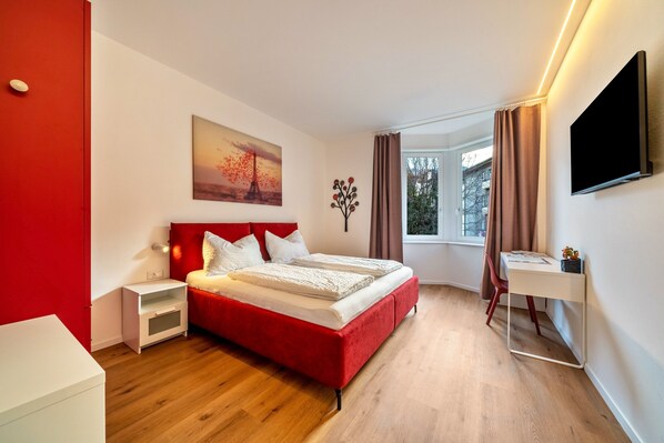 Une chambre d’hôtel moderne avec un lit rouge, un bureau blanc, un téléviseur à écran plat et un tableau représentant un coucher de soleil.