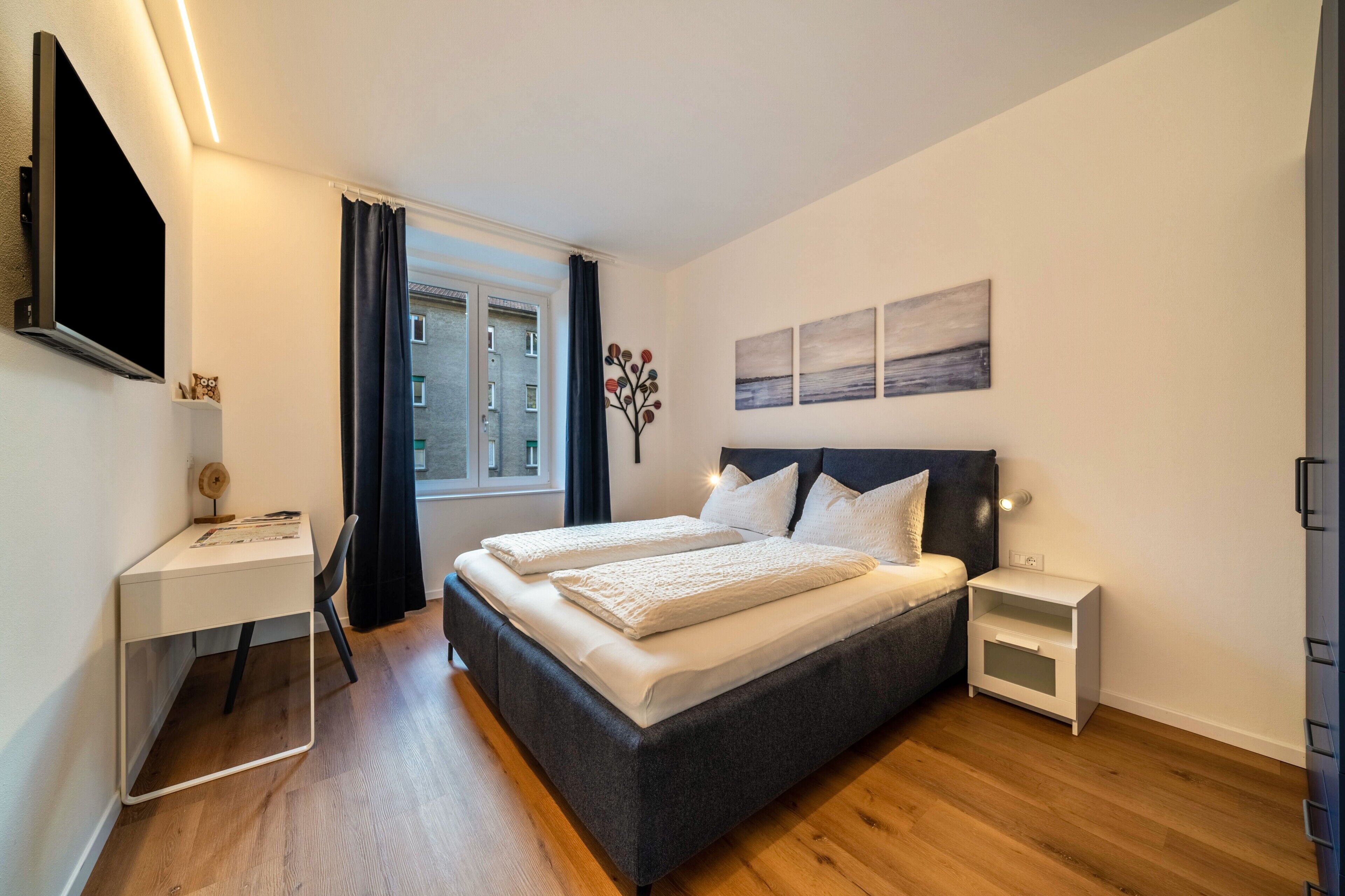 A bedroom with a bed, bedside tables, a desk, a TV, and artwork on the wall.