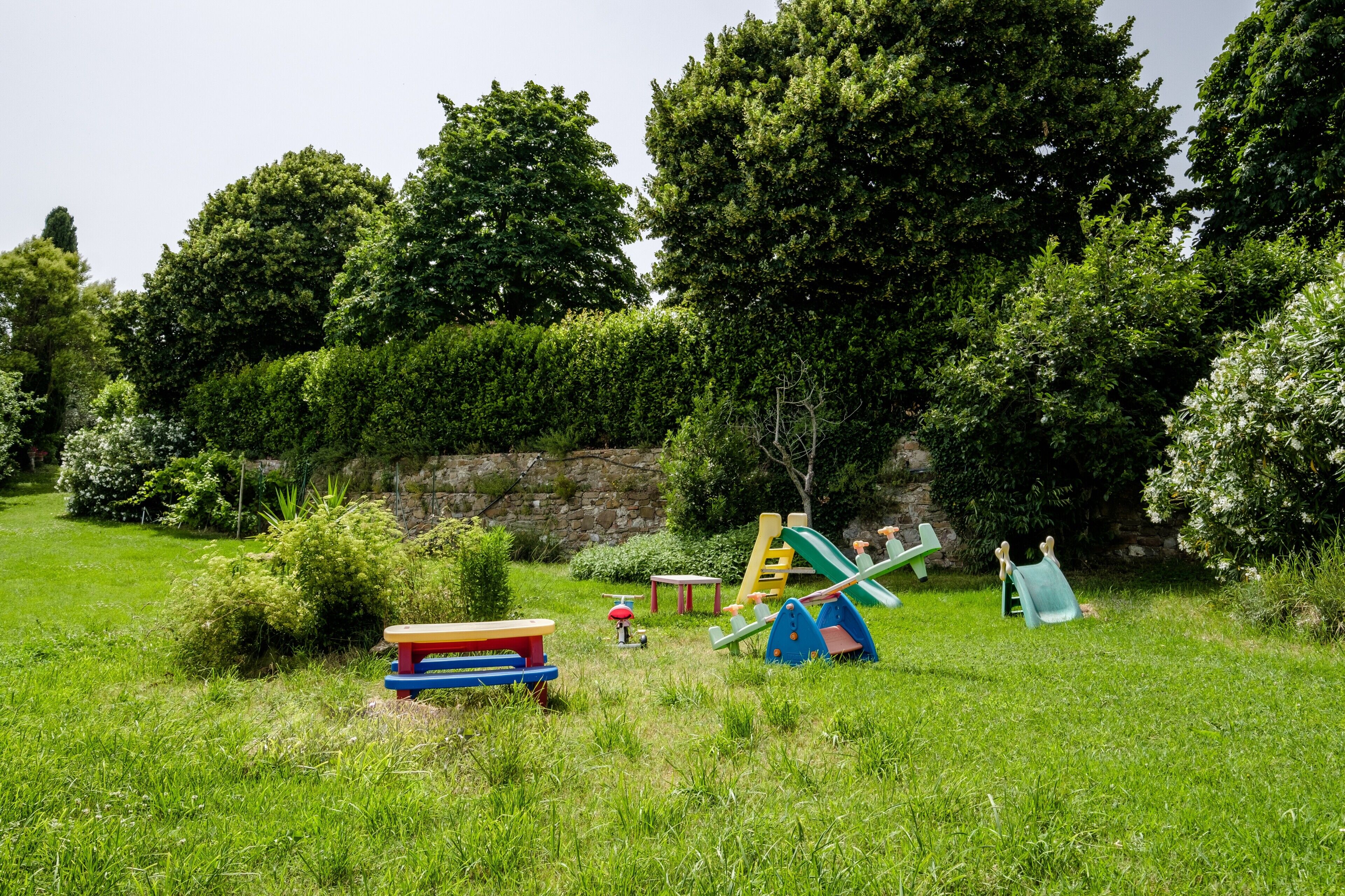 Ein grünes Areal im Freien mit Spielgeräten, einem Picknicktisch und einer Steinmauer im Hintergrund.