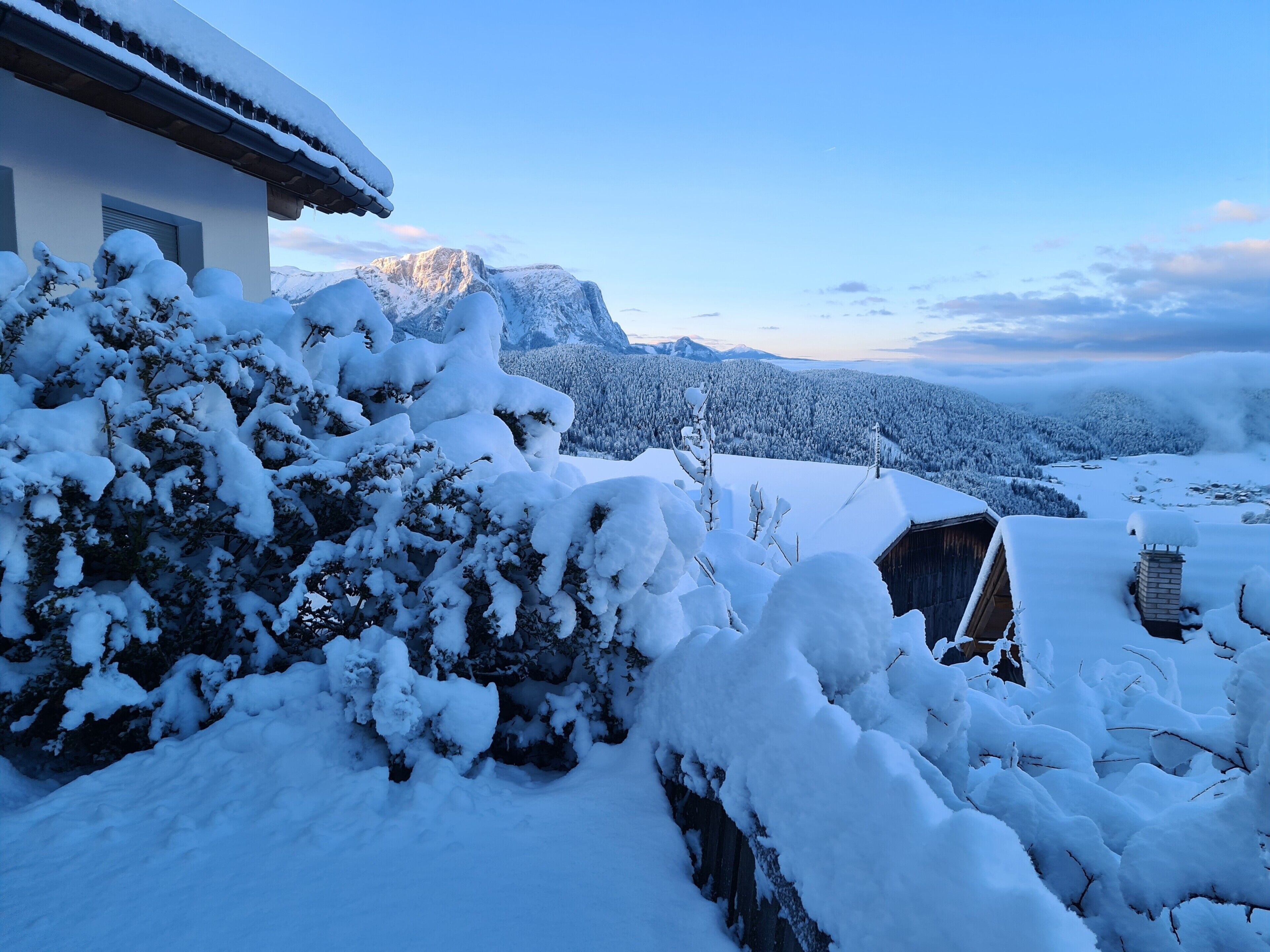 A snow-covered landscape with a house, trees, and mountains in the background.