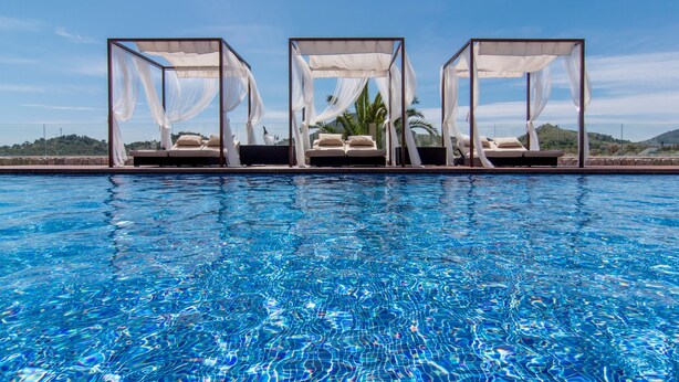 A pool with clear blue water, surrounded by three white canopy beds with white bedding.