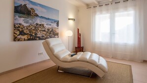 A modern living room with a white leather sofa, a painting of a rocky beach, a floor lamp, and sheer curtains.