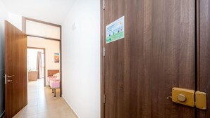 Uma porta de madeira com uma placa, um corredor com chão de azulejos e um quarto com cama e cortinas ao fundo.