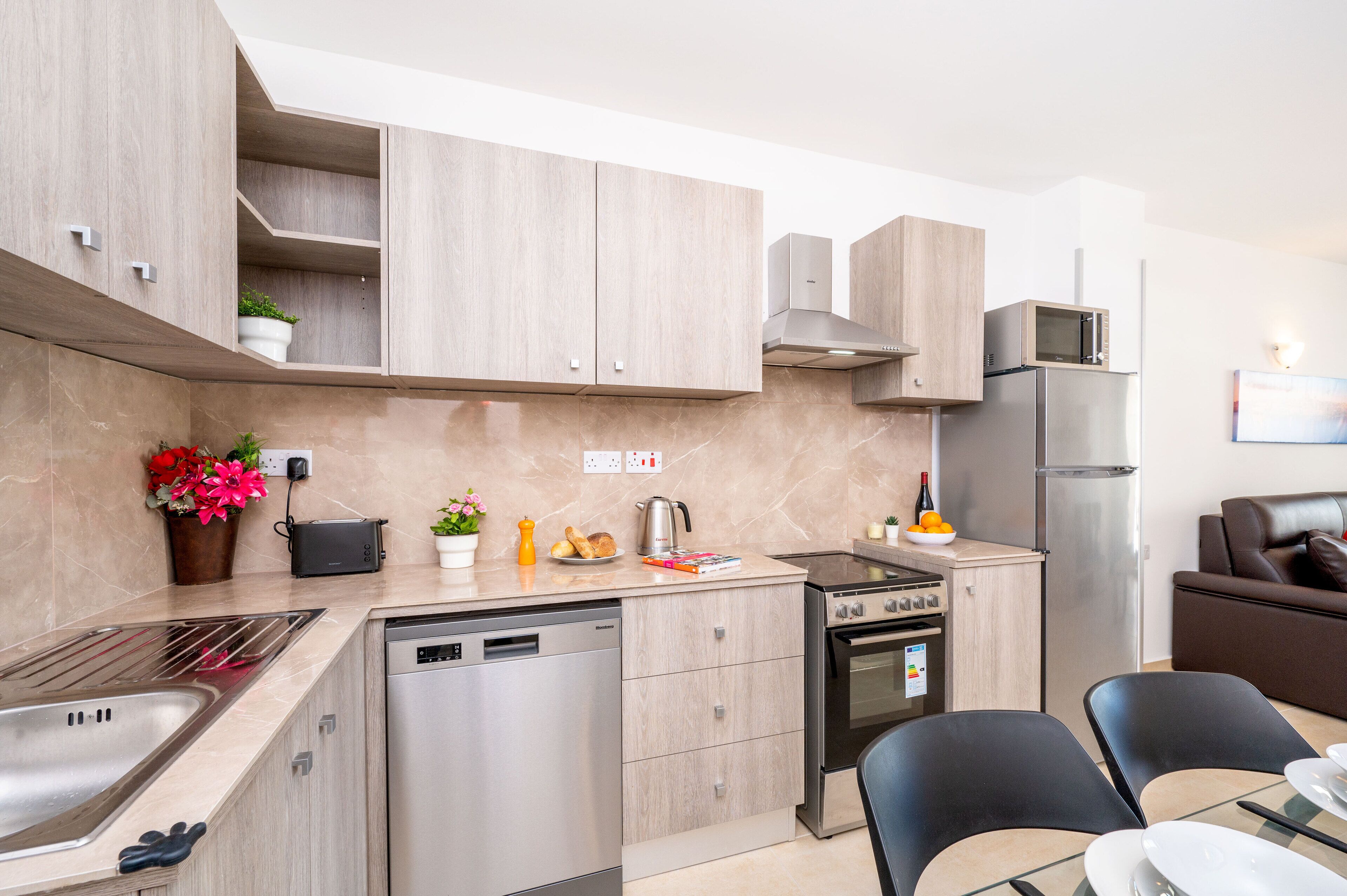 A modern kitchen with light wood cabinets, stainless steel appliances, and a dining area with a glass table and black chairs.
