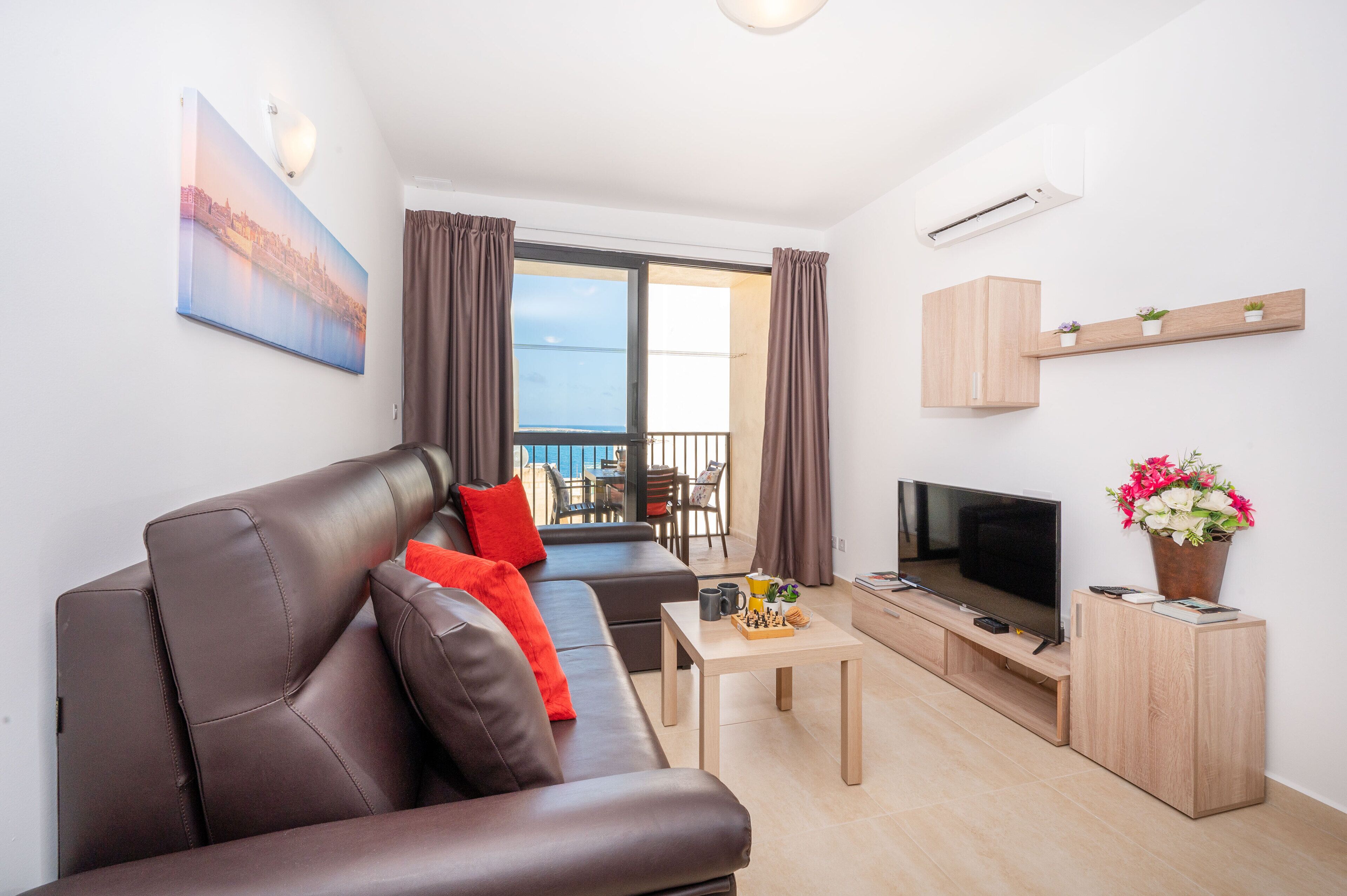 A modern living room with a leather sofa, a flat-screen TV, a coffee table, and a balcony with curtains.
