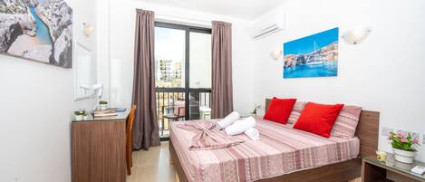 Un dormitorio con cama, escritorio, silla, balcón con vistas y un cuadro en la pared.