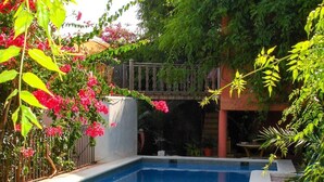 A swimming pool with a tiled deck, surrounded by lush greenery and a red building in the background. - Apartment 'Navila Numero 2' with Shared Pool, Wi-Fi and Air Conditioning (Eivissa)