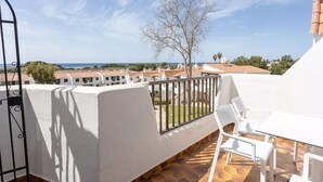 Un balcón con mesa y sillas blancas, vista al mar y edificios con tejados rojos.