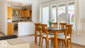 A dining area with a wooden table and chairs, a kitchen in the background, and a view of the outdoors through large windows. - Villa PONTE VECCHIO by Abahana Villas (Teulada)