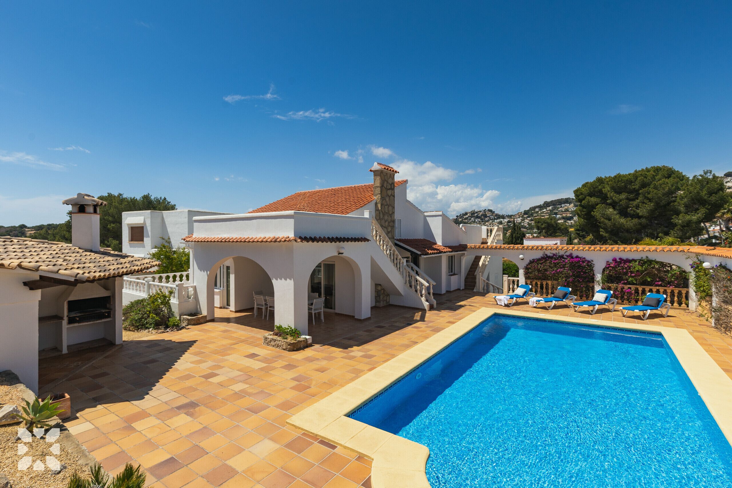 Zona de piscina amb una piscina blava cristal·lina, gandules i un edifici blanc amb teulada d'estil mosaic.