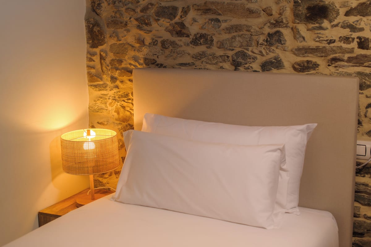 Un lit avec du linge de lit blanc et un seul oreiller appuyé contre un mur de pierre.