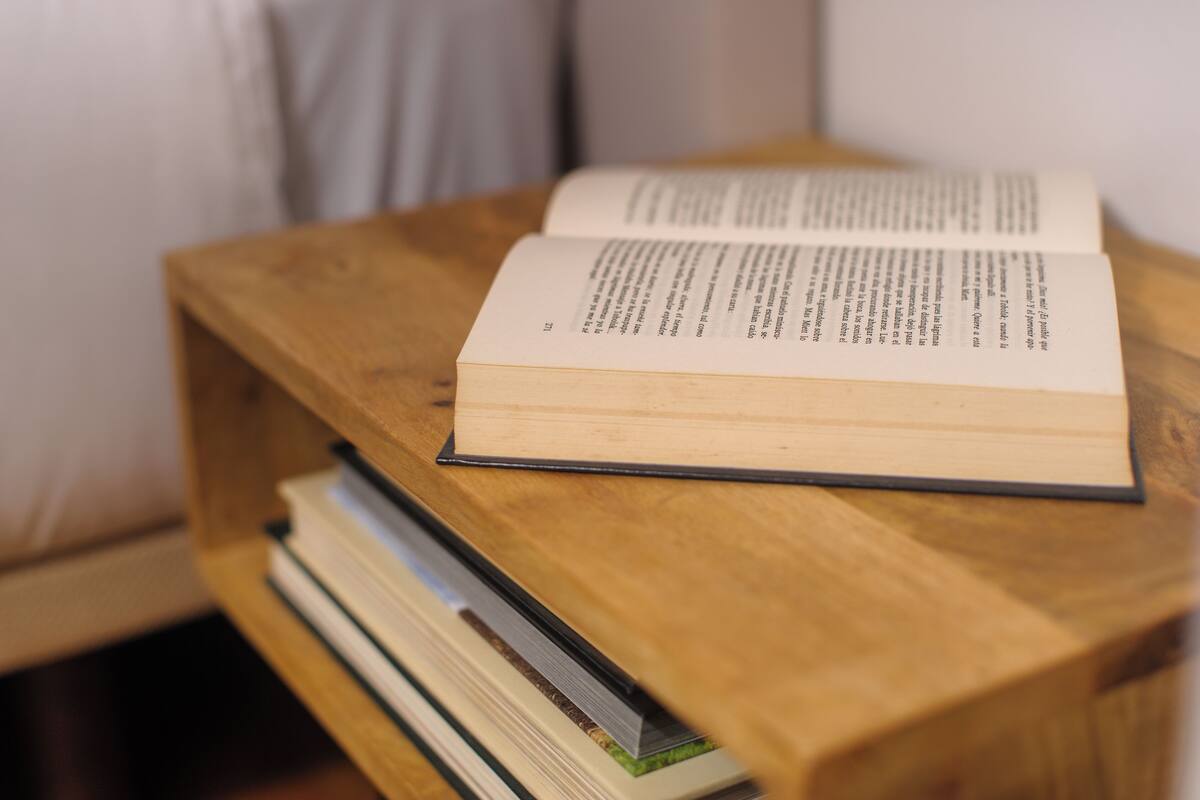 Un livre ouvert est posé sur une petite table en bois, et une pile d’autres livres se trouve en dessous.