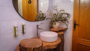 Een badkamer met een rond wasbakje op een houten onderstel, een spiegel, een potplant en twee toiletartikelen op de muur.