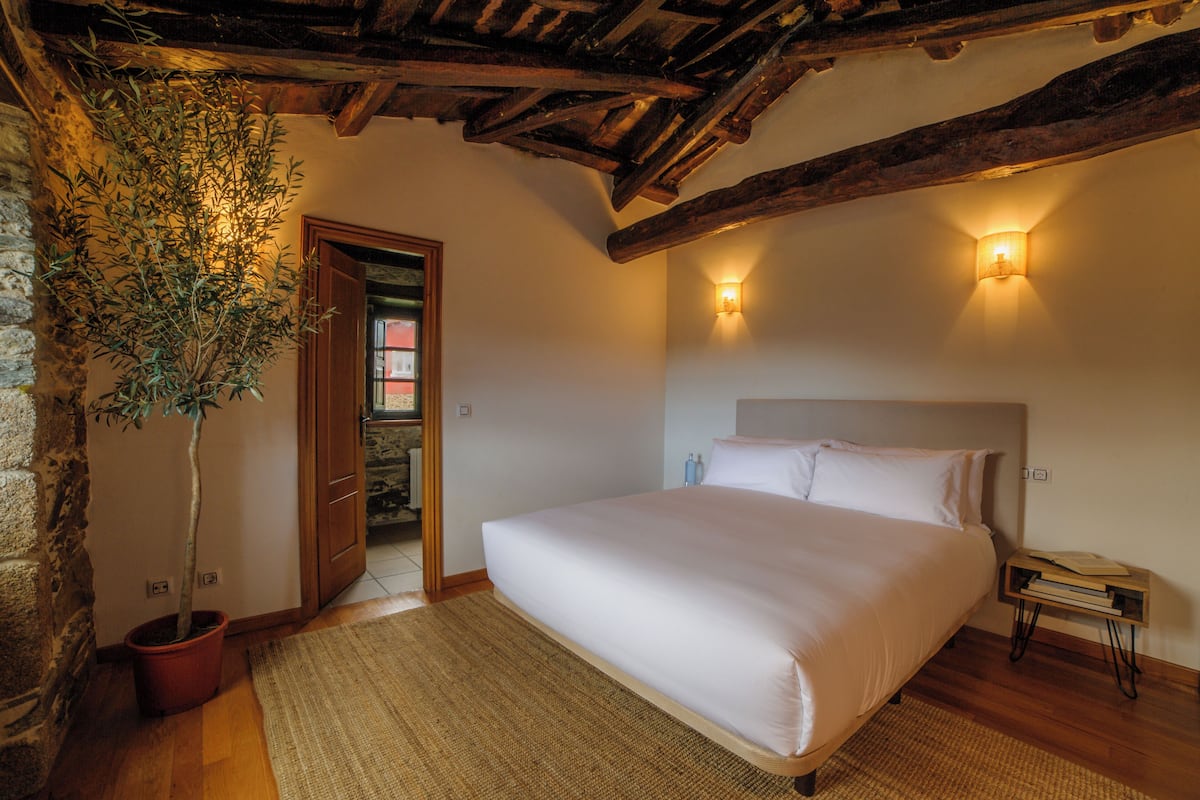 Une chambre avec un grand lit, des poutres apparentes, une plante en pot et deux lampes fixées au mur.