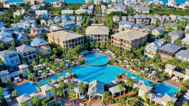 Eine Luftaufnahme eines Resorts mit mehreren Gebäuden, einem zentralen Pool mit Springbrunnen und umliegenden Palmen.