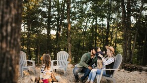 A family sitting on Adirondack chairs in a forested area with a pile of firewood nearby. - Luxury Glamping tent in Ouachita Mountains (Mena)