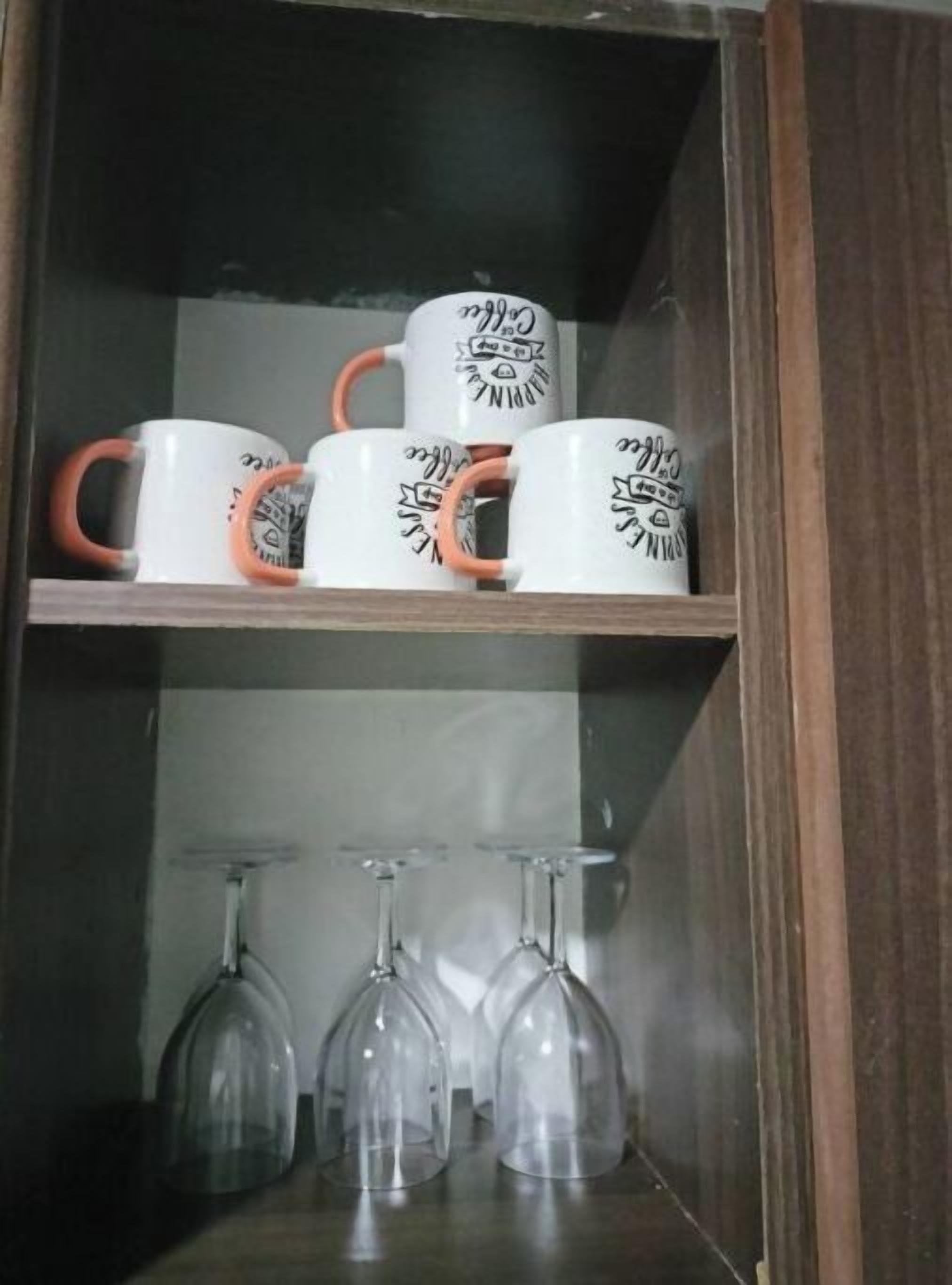 Un mueble de madera con tazas blancas que tienen diseños negros y agarraderas naranjas, y dos copas de vino transparentes en el estante inferior.