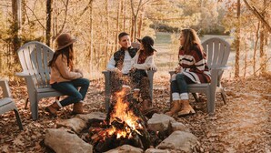 Un grupo de personas sentadas alrededor de una fogata en un área boscosa, con sillas Adirondack.