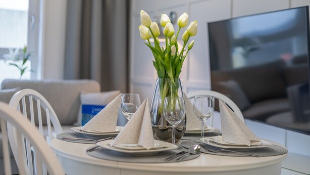A dining table set with white napkins, clear glassware, and a vase with white flowers.