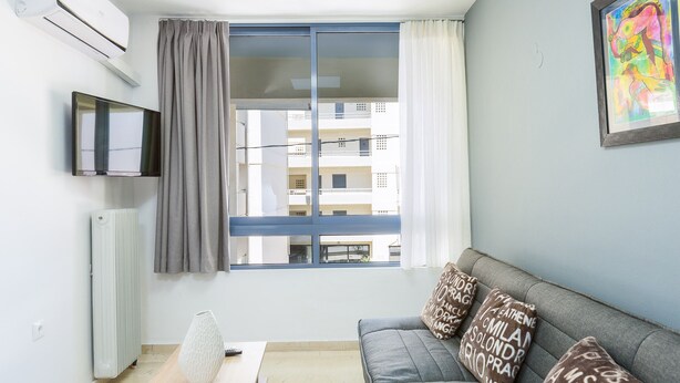 A modern living room with a grey sofa, a flat-screen TV, a radiator, and a window with curtains. - Cozy apartment in the heart of Rethymno town, nr. 1 (Rethymno)