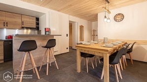 A modern kitchen with wooden cabinets, a dining area with a wooden table and black chairs, and a wall clock. - Le Temps Suspendu "Chardon Bleu" - Relaxation Stay - Apartment 8 people (TERMIGNON)