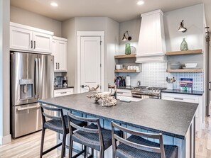 A modern kitchen with stainless steel appliances, a central island with chairs, and decorative items. - Carlton Landing- Wrigley Retreat (Carlton Landing)