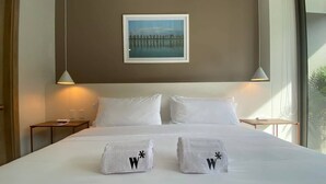 Quarto de hotel com uma cama grande, duas mesas de cabeceira, um quadro na parede e uma janela com vista.