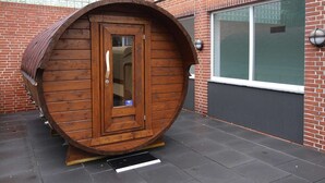 A wooden barrel-shaped structure with a door, positioned outdoors against a brick wall with a window. - Appartement in Westerland auf Sylt (Westerland)