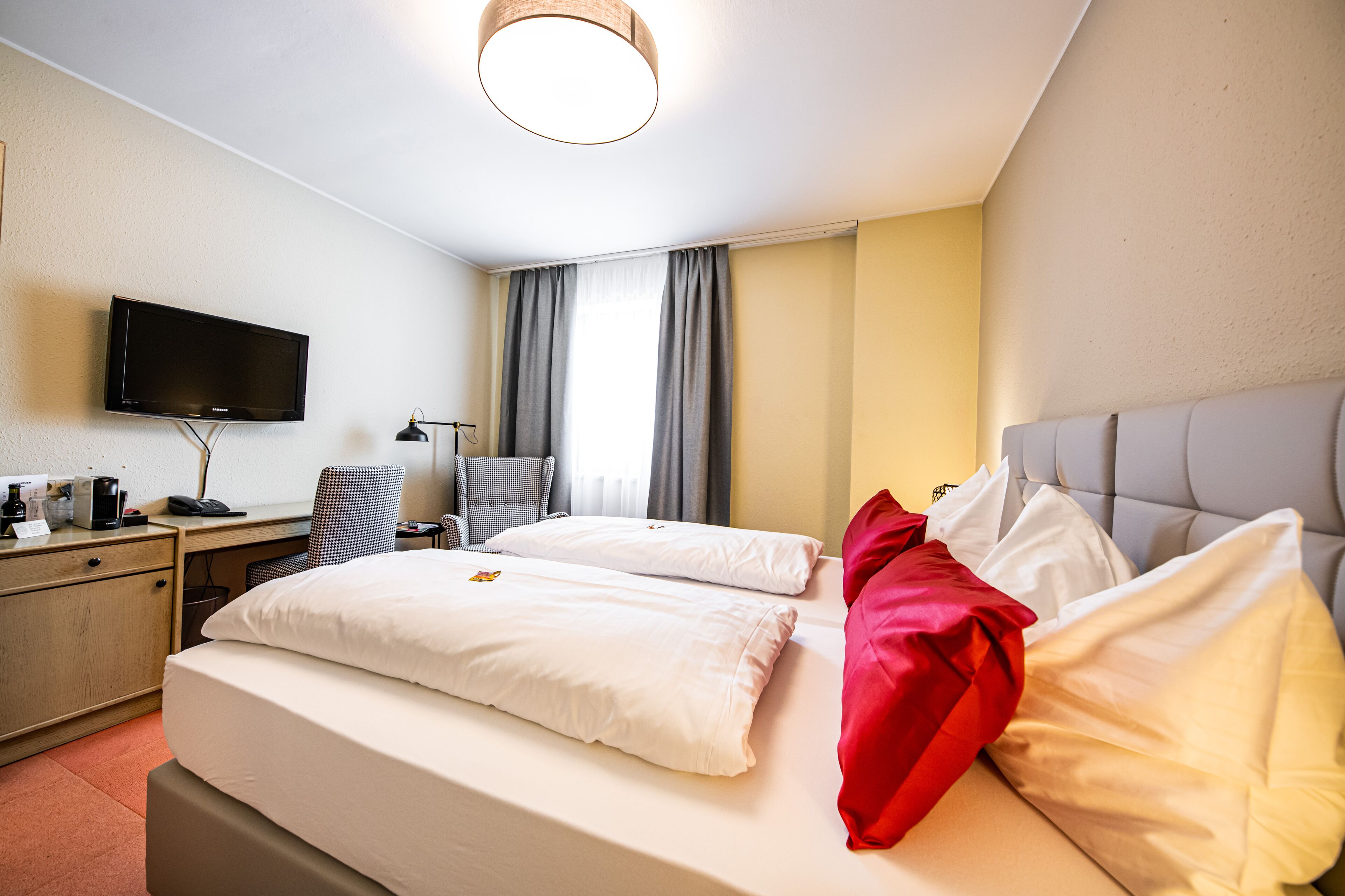 A hotel room with a bed, a desk with a computer, a chair, a television, and a window with curtains.