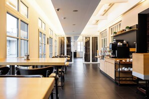 A modern cafe with long tables and chairs, a coffee machine, and shelves with cups and pastries.