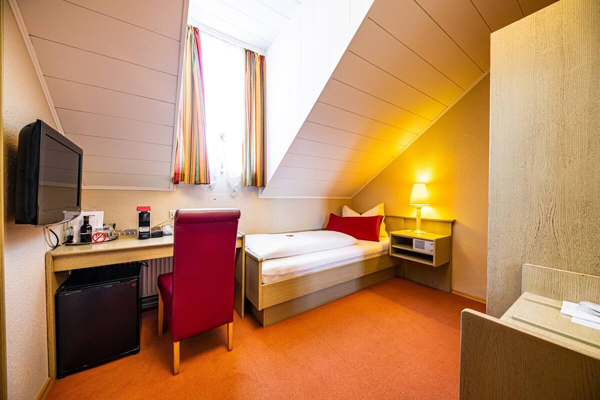 A compact hotel room with a bed, desk, chair, and a small refrigerator.