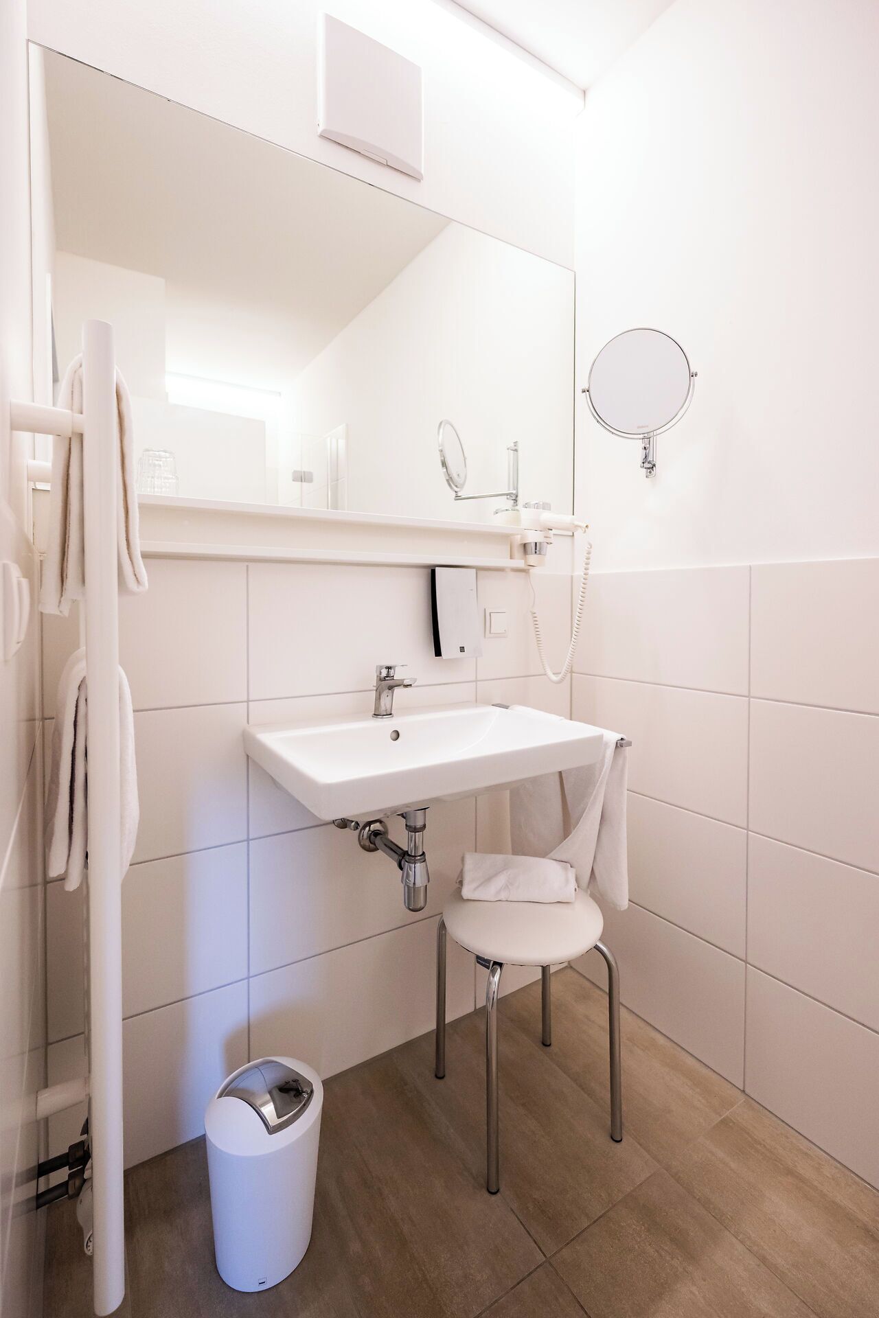 A modern bathroom with a white sink, a mirror, a towel rack, and a trash bin.