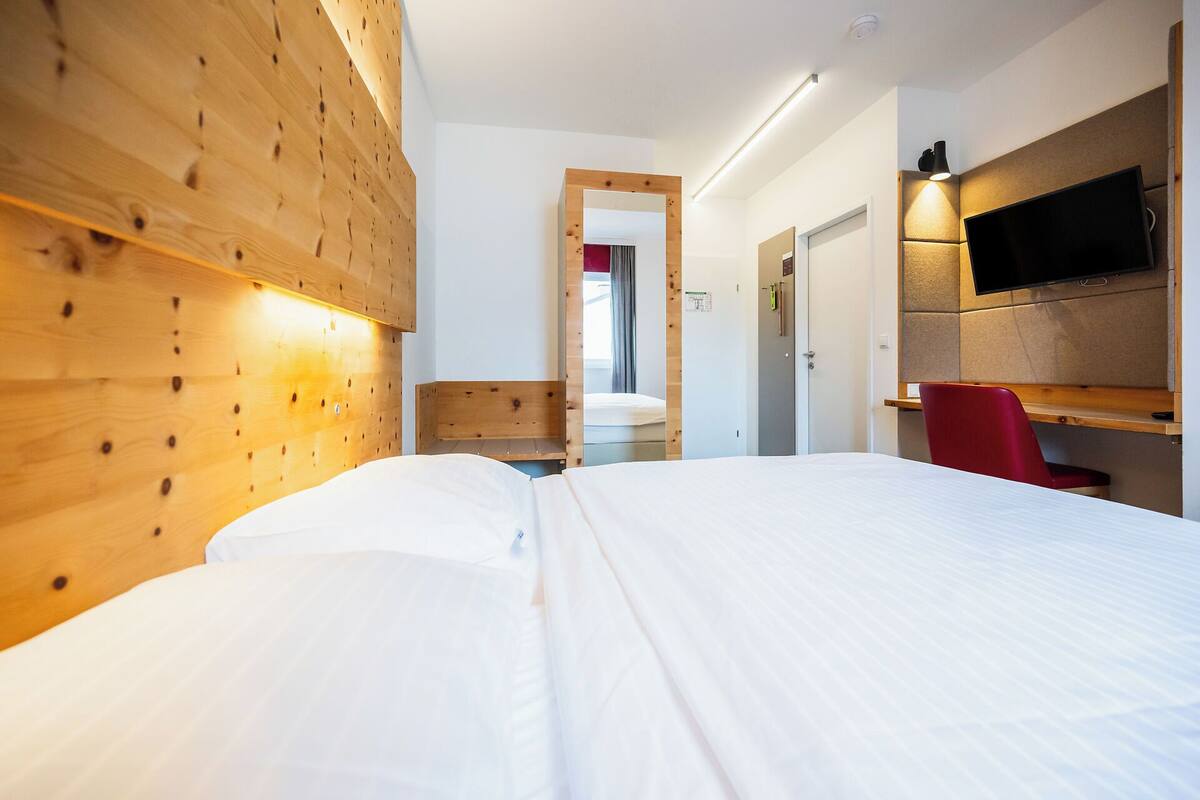A hotel room with a wooden headboard, a bed with white linens, a desk with a chair, a television mounted on the wall, and a door leading to another room.