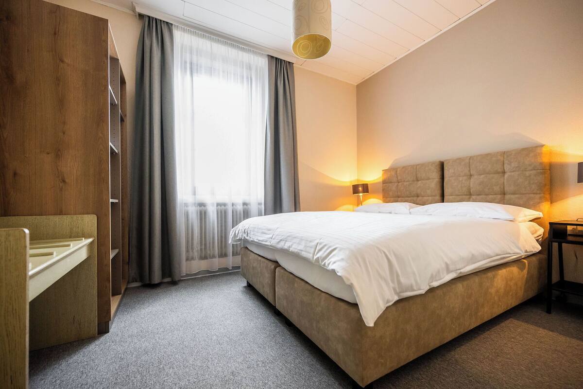 A hotel room with a bed, bedside lamps, a desk, and a window with curtains.