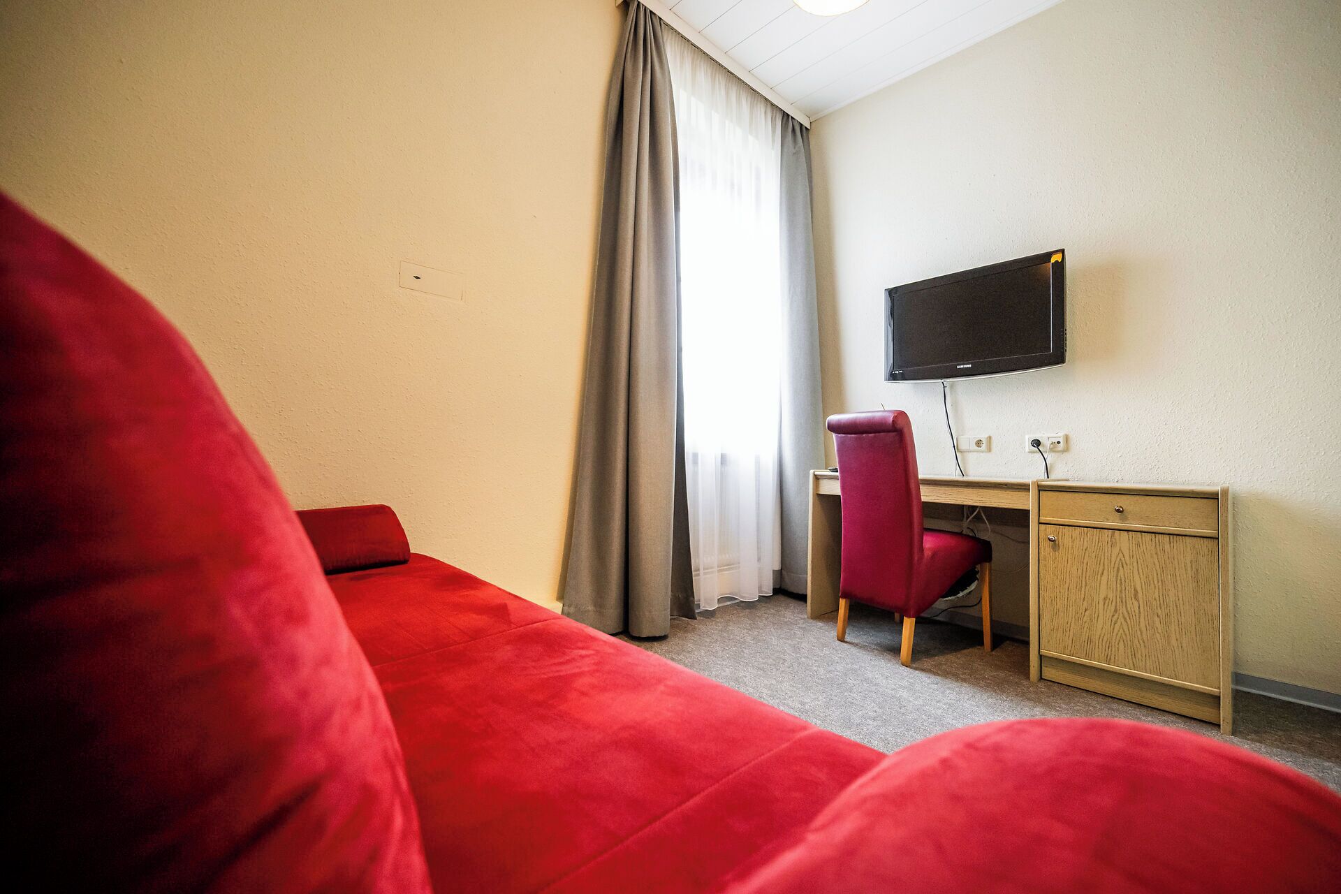 Una habitación de hotel con un sofá rojo, un escritorio con una silla, un televisor en la pared y una ventana con cortinas.