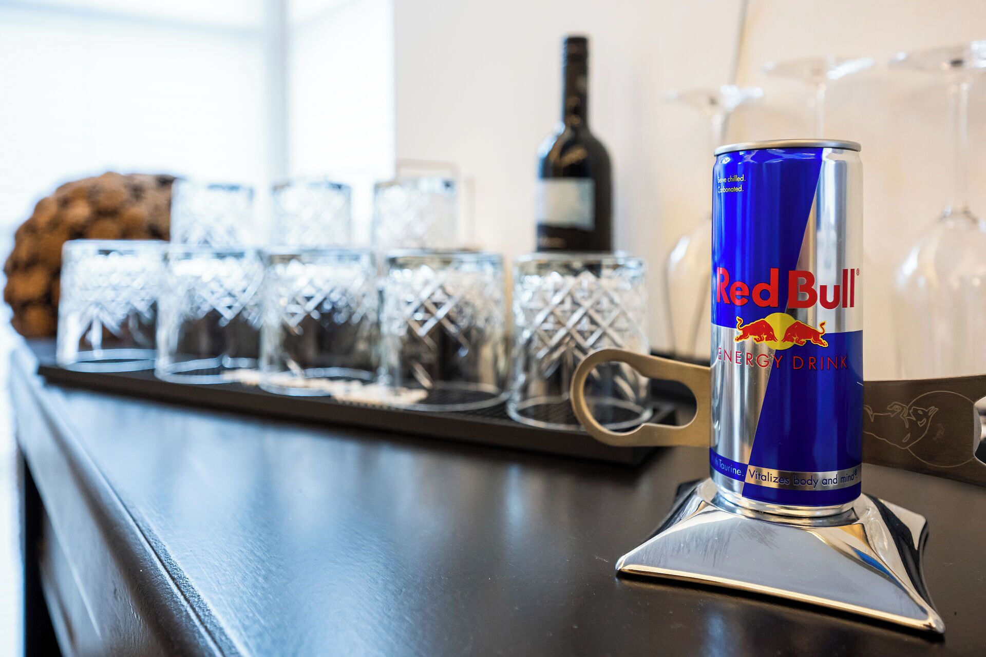 A can of Red Bull energy drink on a bar counter with several empty glasses and a bottle of wine in the background.