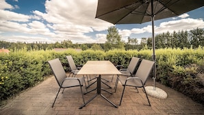 An outdoor patio with a table and chairs, surrounded by greenery and a clear sky.
